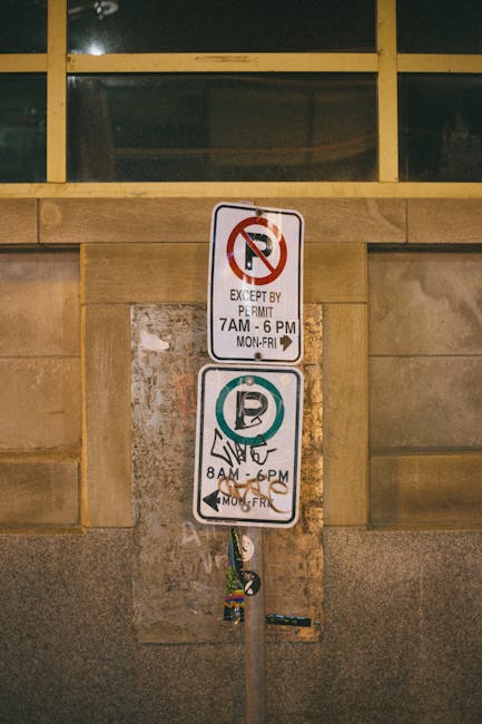 There are two parking signs mounted on a metal pole against a beige stone wall. The top sign indicates no parking except by permit from 7 am to 6 pm, Monday to Friday, with a red circle and slash over