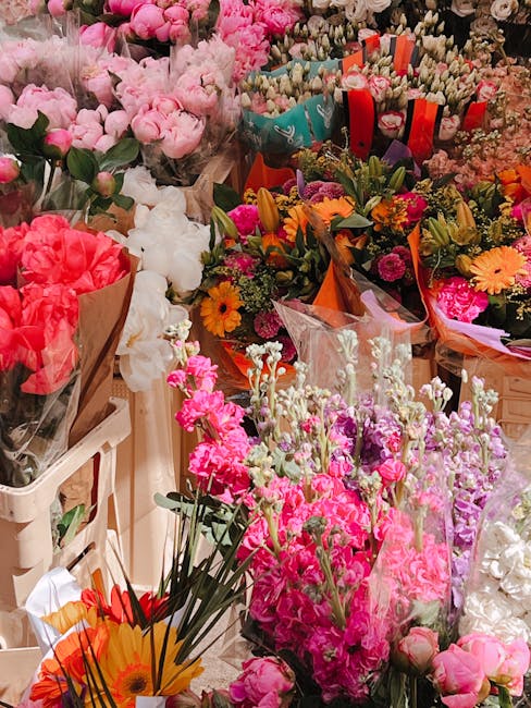 A vibrant display of fresh flowers arranged in various bouquets and wraps, featuring a mix of pink, white, orange, and purple blooms. The arrangement includes peonies, daisies, and other seasonal flow