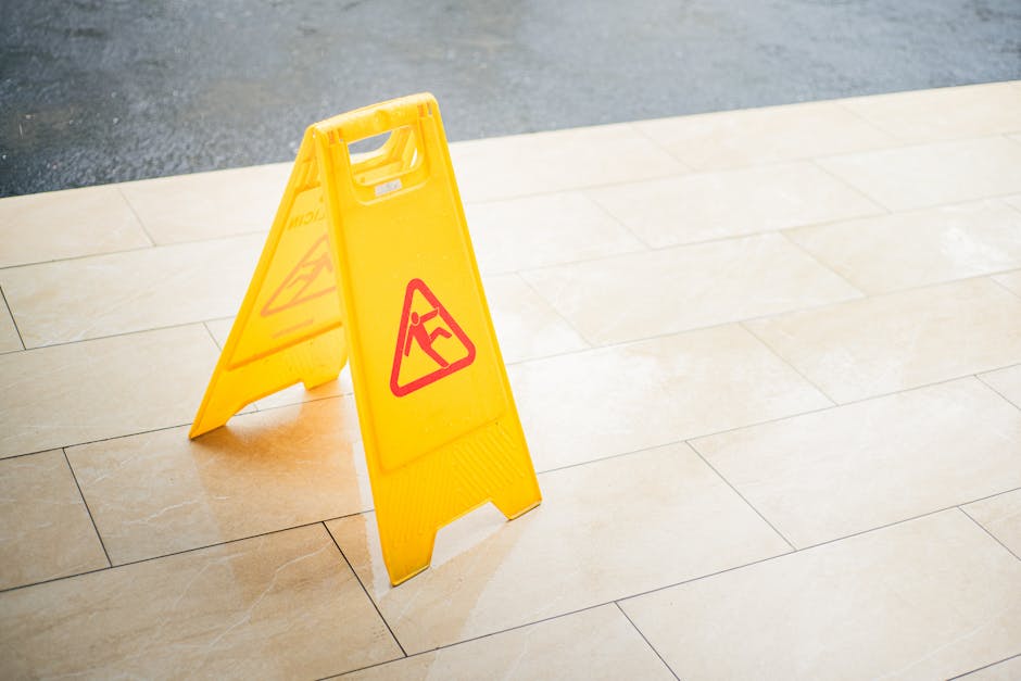 A yellow, foldable wet floor warning sign with red triangular caution symbols is placed on a light beige tiled floor near an outdoor paved area. The sign is positioned upright, alerting passersby to p