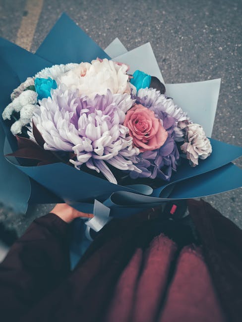 A vibrant floral bouquet held by a person, featuring a mix of fresh flowers including a large white chrysanthemum, soft pink roses, lavender-hued lisianthus, and small turquoise accent flowers. The bo