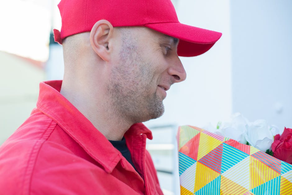 A man dressed in a red cap and a matching red jacket holding a floral gift wrapped in colorful, geometric-patterned paper with yellow, blue, red, and white shades. The bouquet contains white and red f