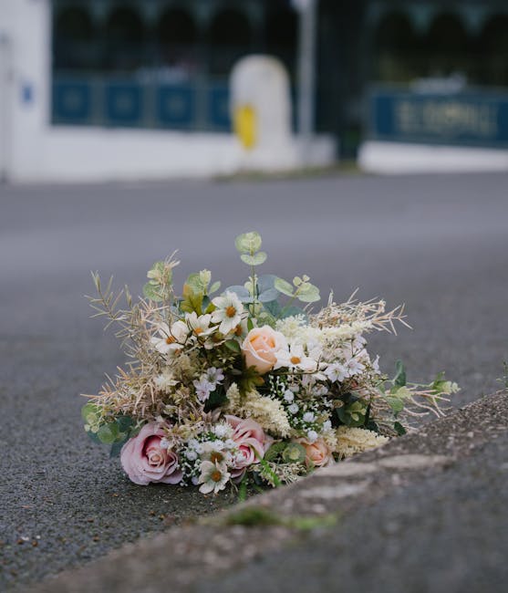 A delicate floral bouquet placed on an urban street surface, featuring soft pink and white roses, small white daisies, and sprigs of greenery including eucalyptus and thyme, arranged with a light, air